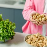 Hands presenting peanuts with a bowl, lettuce, avocado, and tomato nearby on kitchen counter.